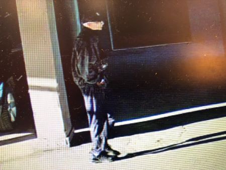A right side photo of the described suspect walking past vehicles in a parking enclosure. 