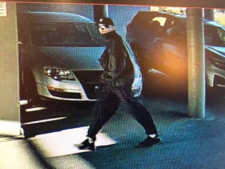 A left side photo of the described suspect walking past vehicles in a parking enclosure. 