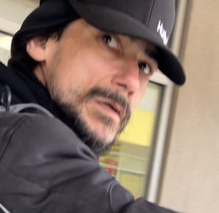 A photo of a suspect looking over his right shoulder. He is wearing a black jacket, ball cap, and has facial hair. 