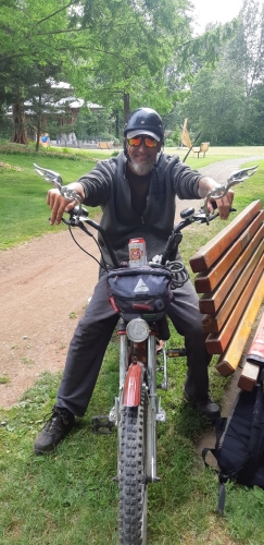 David Phillips, in a park, sitting on a red e-bike with Chopper style handlebars. He has a grey beard and is wearing sunglasses, a black helmet and dark clothing
