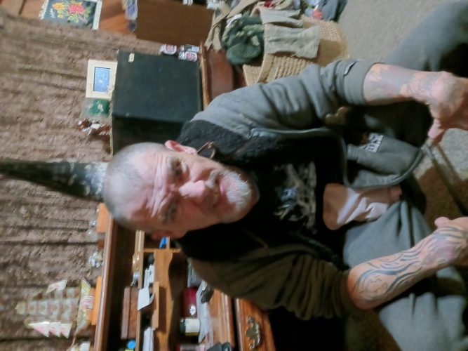 David Phillips, sitting down wearing an open hoodie, sleeves rolled up to show his tattooed arms. He has a grey beard and very short grey hair