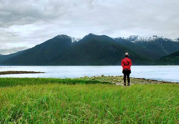 RCMP officer in red serge standing in grass facing a large body of water and mountains