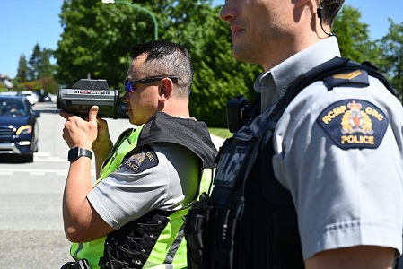 Two Burnaby RCMP officers in uniform stand beside a road on a sunny day with one using a laser speed detector