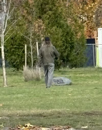 Un homme s’éloigne à pied dans un champ. Il a de longs cheveux bruns grisonnants et porte un chapeau noir, une veste brun pâle ou grise ainsi qu’un pantalon gris.