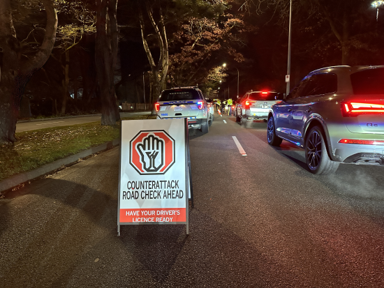 pop-up sign on the road that reads <q>CounterAttack road check ahead, have your driver’s licence ready</q> with a police vehicle and cars in the background