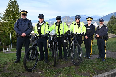 Six policiers portant divers uniformes se trouvent sur une zone herbeuse avec des montagnes en arrière-plan. Trois des agents ont des vélos électriques devant eux.