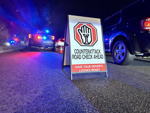 Sign that reads: CounterAttack road check ahead. Have your driver’s licence ready.</q> With cars in the background.