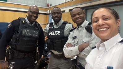 Le gend. Dwight Martin se tient en souriant aux côtés de trois autres policiers dans un gymnase lors d’une activité menée dans le cadre de la Journée de l’excellence des Noirs à Cloverdale Traditional School (de gauche à droite : gend. Dwight Martin, cap. George Amoako, gend. Berthier Kyobela et insp. Veronica Fox)