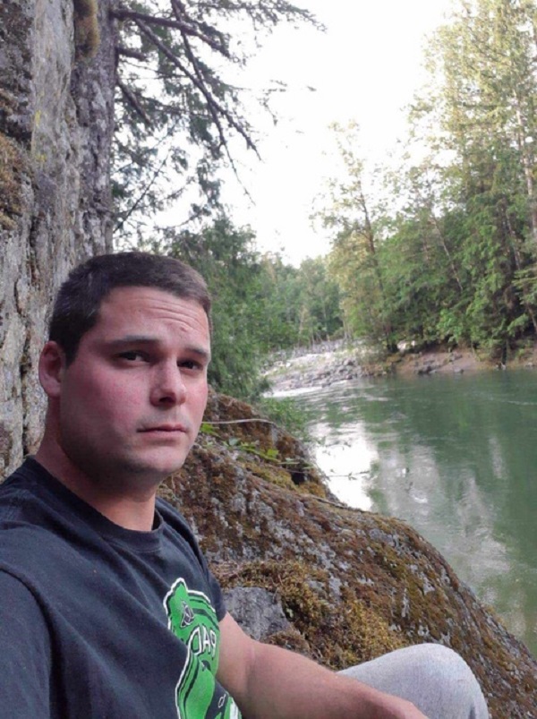 First Nations male wearing dark t-shirt with green logo sitting outside beside a river