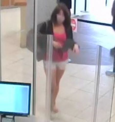 A Caucasian woman in a black blazer, pink mask, and white shorts with shoulder length brown hair walks into a commercial business with white tile floors.