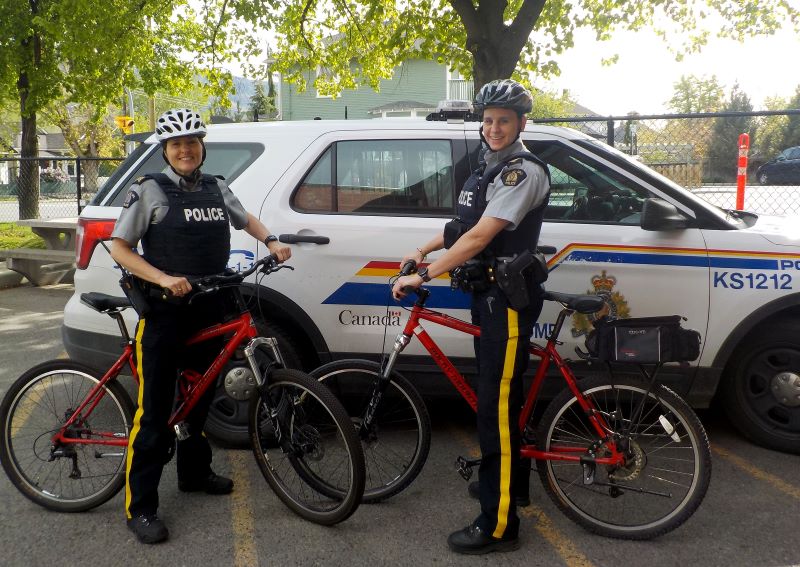 Two uniformed police officers (fem) wearing bike helmets are on red police bikes in a parking lot. A white police SUC is behind them.  