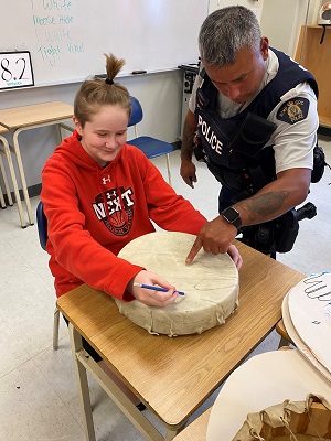 Photo of Cst. Williams standing at a classroom desk, showing a student how to build a circular drum.