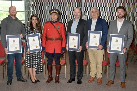 Civilians in business wear stand holding framed RCMP appreciation awards. An  RCMP Officer in Charge is wearing his red serge in the middle.