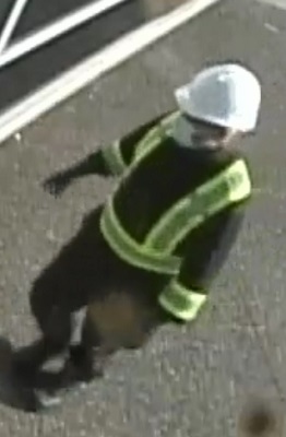 suspect no 1 portant un casque blanc, un masque, un chandail noir avec un gilet de haute visibilité, un pantalon noir, des gants noirs et des bottes noires.