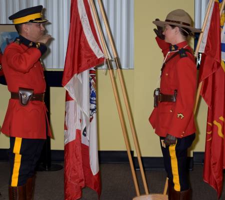 Supt. Syd Lecky presents Cst. Crystal Evelyn to the rank of corporal during a ceremony earlier this summer. On the left, a man in red serge salutes a female member is red serge. Canadian and BC flags are in the background. 