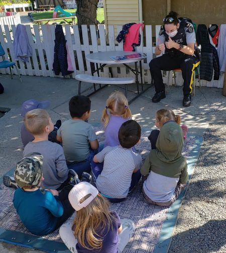 : A photo of Cpl. Napier in one safety discussions with kids. (Kids sit in a group in front of a uniformed police woman who is giving them the thumbs up. Children coats hang on a white wooden fence behind her. 