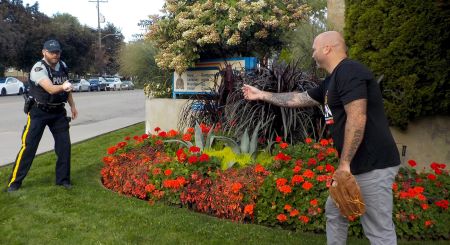From left, a man in a police uniform plays catch with a man in a black t-shift and slacks, on the grass in front of a Kamloops RCMP detachment sign, surrounded by flowers and landscaping.