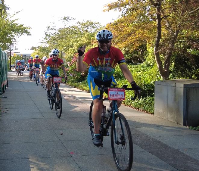 A group of smiling Cops for Cancer cyclists approach Burnaby City Hall on their bikes. 