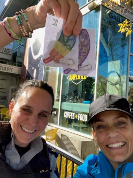 De gauche à droite, Cpl. Dana Napier et Ingrid Brakop, coordonnatrice de la sécurité routière et de la communauté à l’ICBC, tiennent un réflecteur à fermeture à glissière de couleur arc-en-ciel en forme de plume, devant un café du centre-ville avec une écriture jaune à la fenêtre. 