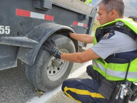 A police officer in a yellow vest crouches down to inspect a trailer tire.