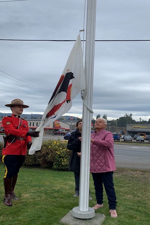 photo of RCMP officer in red serge assisting Okanagan Indian Band elder raising the Okanagan Nation Flag