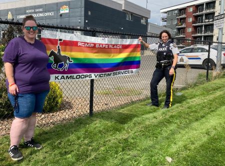 À gauche, Ashton O’Brien de la Kamloops Pride Society, vêtue d’un chandail violet et d’un short en jean, tient une bannière arc-en-ciel avec la caporale de la GRC, Dana Napier. Le drapeau est orné d’une silhouette d’un gendarme à cheval et de la mention « RCMP Safe Place » (Espace sûr de la GRC) en haut et « Kamloops Victim Services » (Services aux victimes de Kamloops) en bas. 