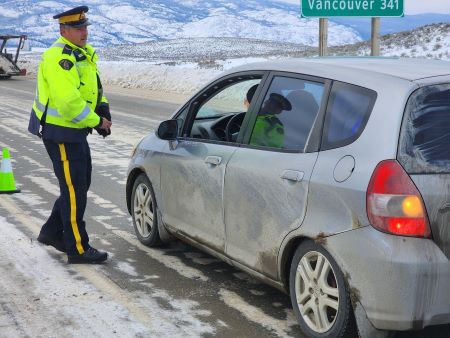 A police officer with a yellow safety vest approaches a grey car on the side of a snowy highway.