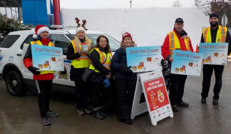 A team of Crime Prevention volunteers and staff, wearing high visibility vests, gathered in front of a police cruiser with signs for Stuff the Cruiser.