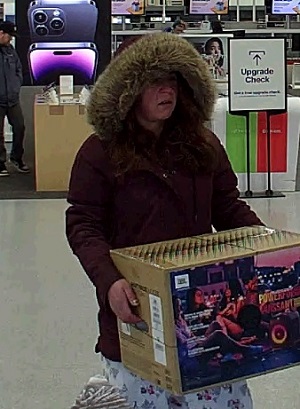  Femme quittant le magasin Best Buy avec une boîte de haut-parleur. Elle porte un parka rouge avec une capuche en fourrure relevée et un pantalon à carreaux blanc.
