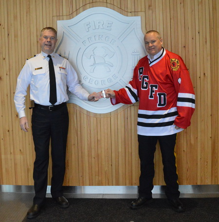 Photo du chef de pompier Warner et du surintendant Wright de la GRC à la caserne de pompier 1. Le surintendant Wright porte le chandail de hockey de l’équipe des pompiers de Prince George et remet un billet de 50 $ au chef de pompier Warner.  