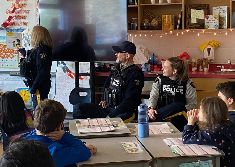 One Youth RCMP officer in uniform reading a book to a class of elementary school students while two uniform youth RCMP officers sit nearby