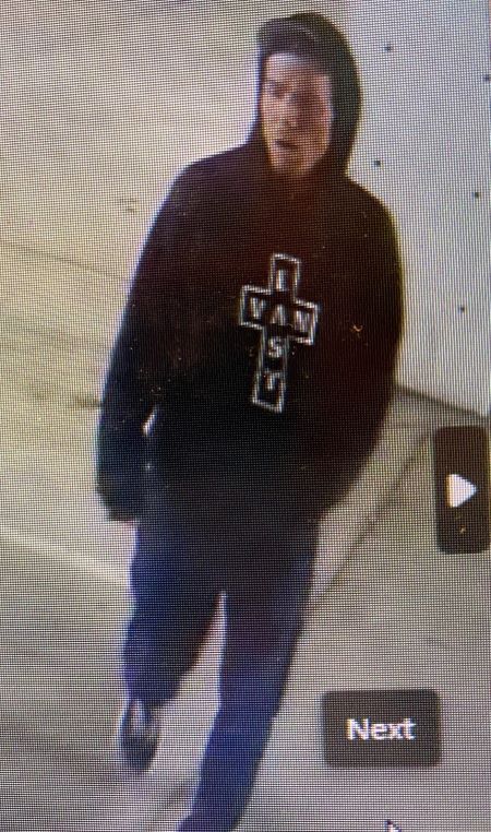 A Caucasian man wearing a black hoodie sweater with an image of a cross containing white writing inside, blue pants, and black sneakers with white laces walks through a parking garage.