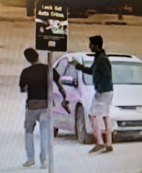 Deux hommes d’origine sud asiatique vêtus de hauts foncés se tiennent debout devant une voiture grise. L’un d’entre eux tient une arme de poing potentielle.