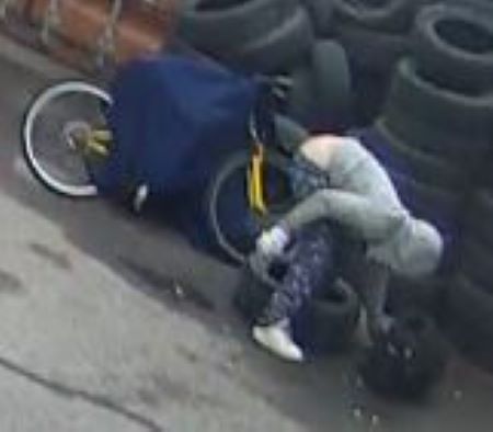 Suspect leaning over: A person in a grey hoodie and blue pants leans over in front of a tire pile and tends to a black bag. His pants are falling down slightly. A green and yellow bike is beside him with a blue blanket draped over top. A white bulky glove or cast is on his right hand and forearm. 