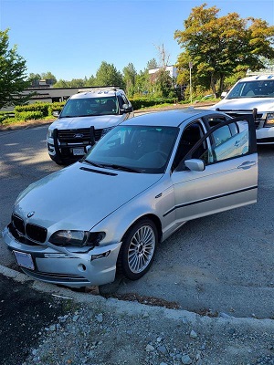 Photo of grey BMW with front-end damage, parked with two RCMP vehicles behind it