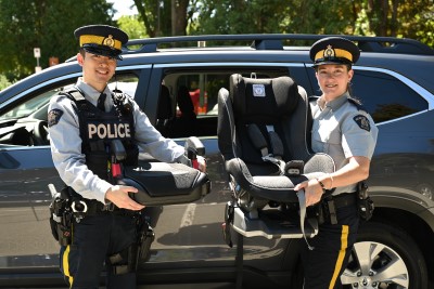 Two smiling police officers hold a car seat and booster seat beside a vehicle outdoors