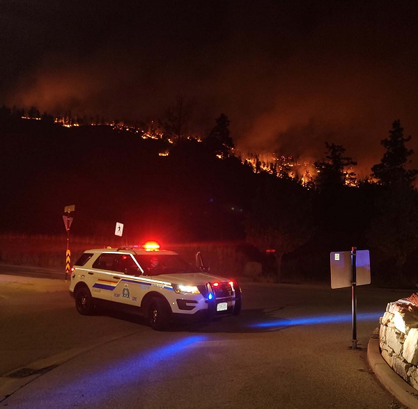 Police vehicle at an intersection with emergency lights on and wildfire in the background at night