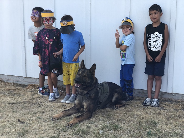 •&#9;Cinq jeunes garçons avec de la peinture faciale et portant des casquettes de police pour enfants sourient à côté d’un gros chien policier brun. 