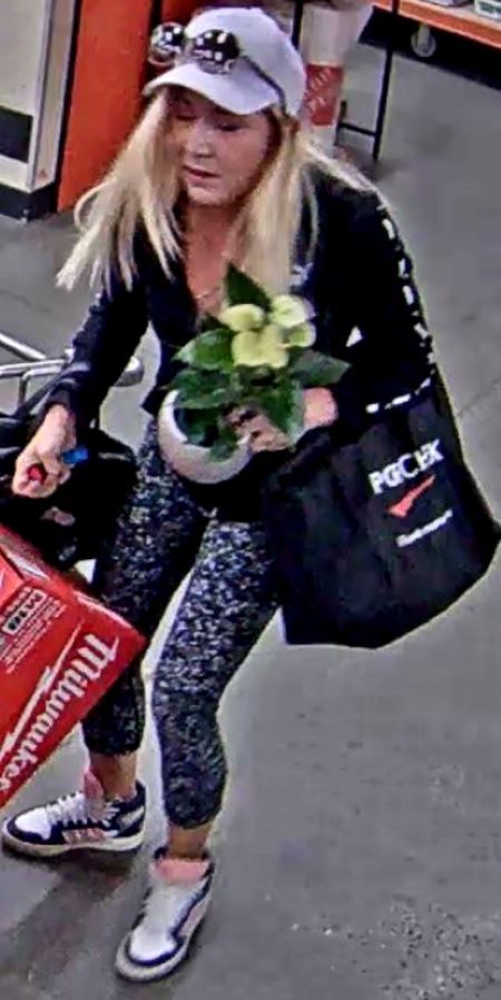 A blond woman wearing a lilac ball cap, wearing floral tights, a black top, and white, black, and pink runners, holding a potted flower and a power too box. 