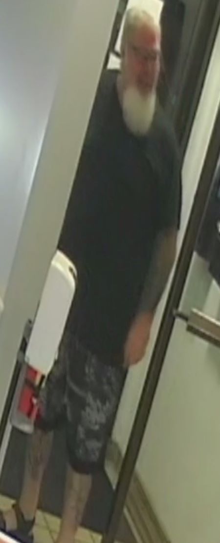 The suspect stands in a doorway. He is a heavy-set Caucasian man with grey hair, white beard, and black framed glasses. He is wearing a dark t-shirt and shorts. 