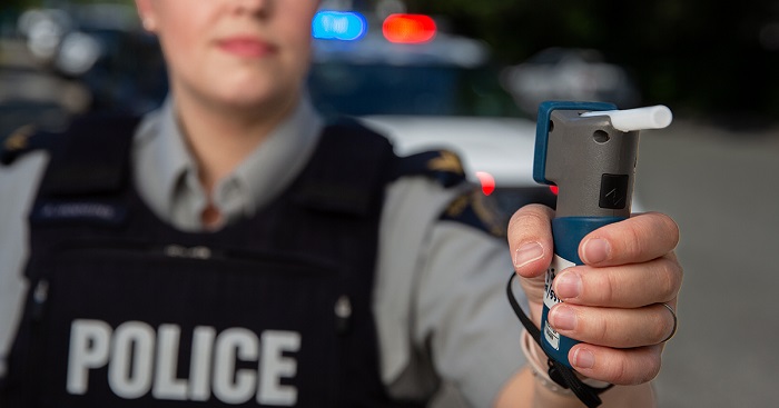 RCMP Police officer holding an Approved Screening Device in front of a marked police car with lights on