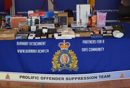Various stolen items laid out on a table with a Burnaby RCMP table cloth with the RCMP logo on it. The logo states <q>Royal Canadian Mounted Police, Maintien Le Droit. The cloth also reads <q>Burnaby Detachment, www.burnaby.rcmp.ca, Partners for a Safe Community</q>. There is also a second banner reads </q>Prolific Offender Suppression Team</q>.
