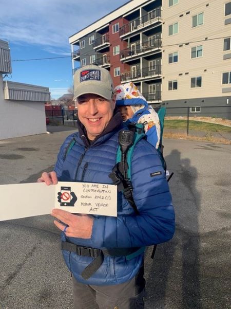 Un homme portant un manteau bleu et un porte-bébé avec une poupée sur son dos tient un carton sur lequel les gens peuvent lire que d’utiliser son téléphone au volant constitue une infraction.   